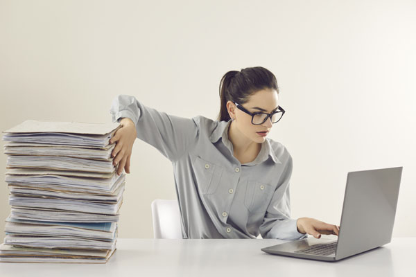A woman working on a computer, sitting at a desk with a pile of papers that she will have to scan.