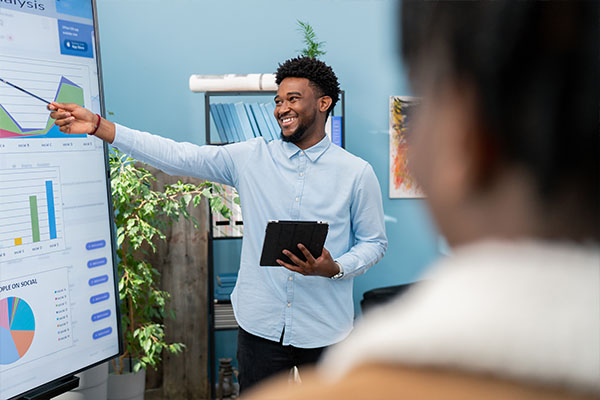 A young man standing with a tablet in his hand showing the results of his study on a large screen during a presentation.