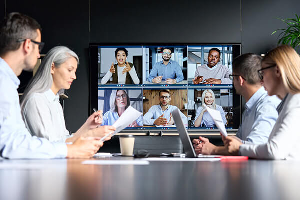 un groupe de personnes autour d’une table qui suivent une vidéoconférence sur un écran géant.