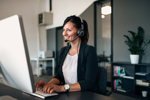 une femme souriante avec une casque d’écoute en conversation téléphonique devant son écran.