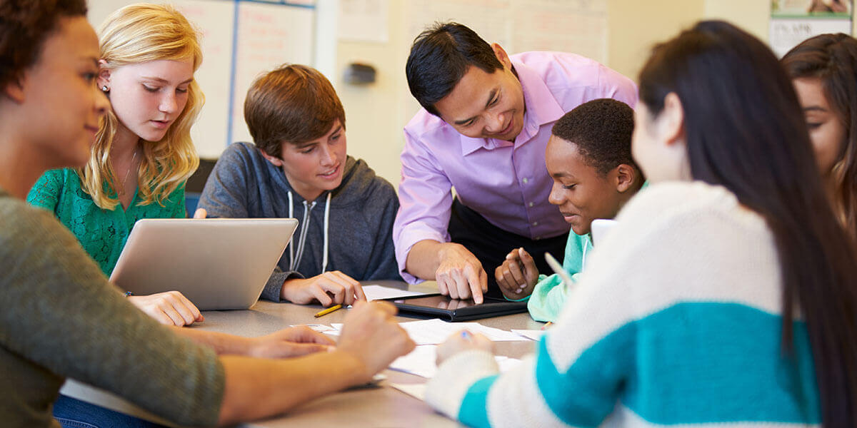 Groupes d’adolescents assis autour d’une table avec un enseignant pointant une tablette électronique.