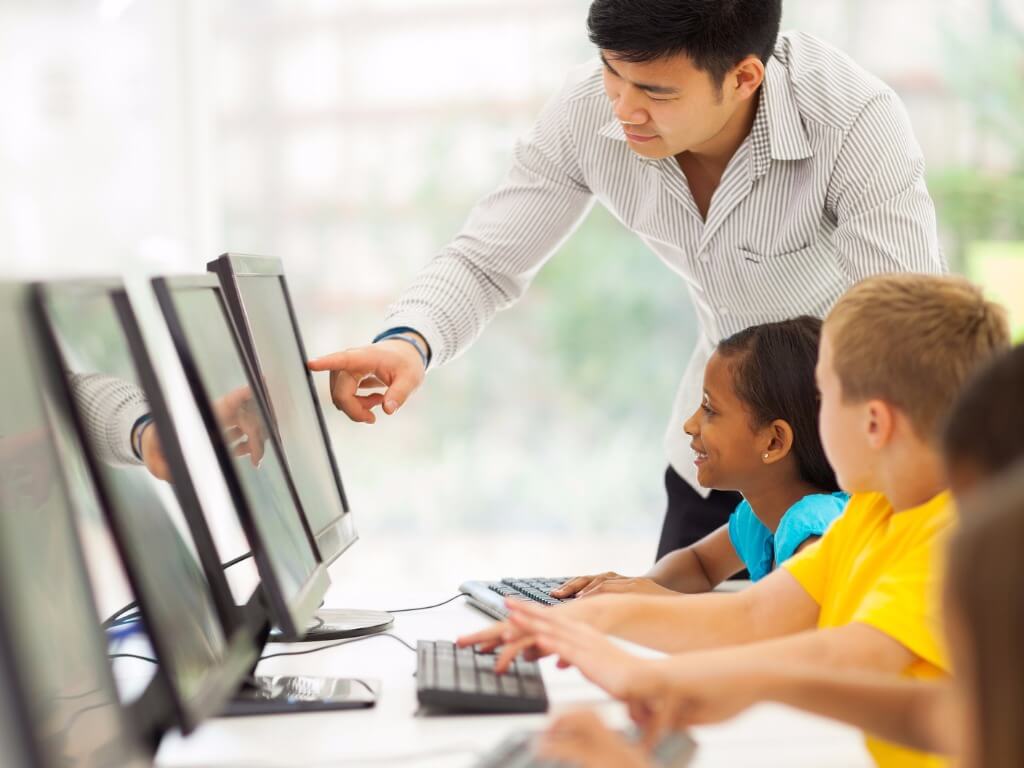 Un enseignant, penché derrière une élève du primaire, l’aide à utiliser un ordinateur pour accéder à des activités d’apprentissage.