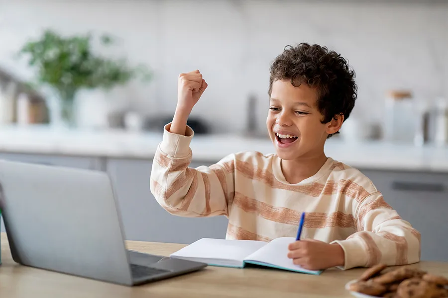 Un enfant célèbre une réussite devant un ordinateur portable tout en prenant des notes dans un cahier.